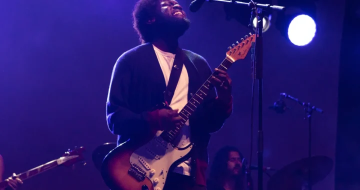 Michael Kiwanuka performing live with a Fender guitar under purple stage lighting