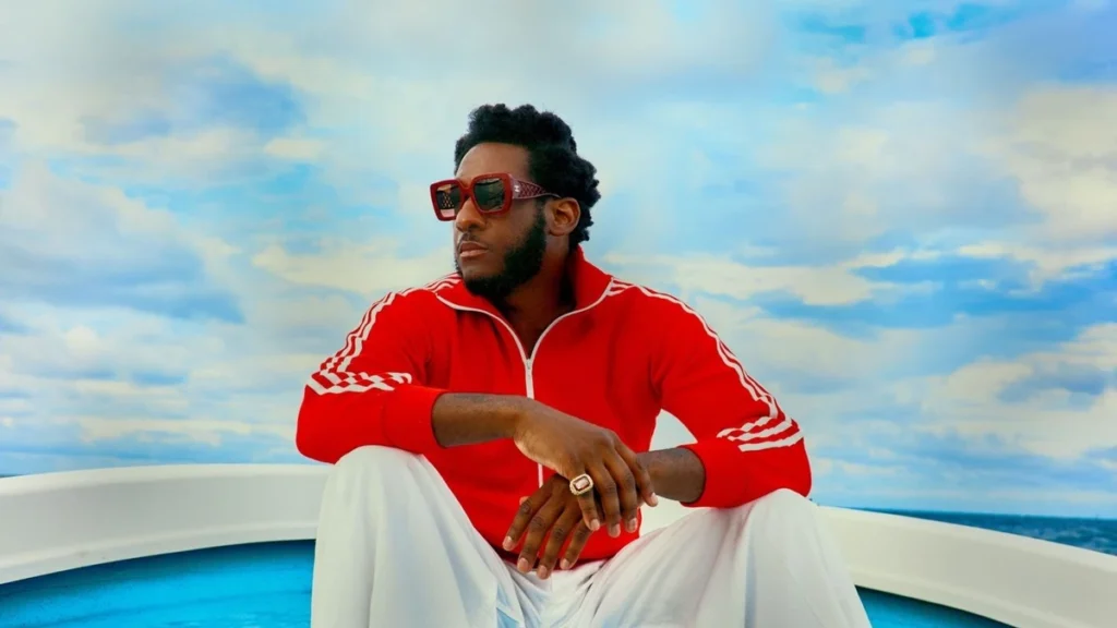 Leon Bridges in a red tracksuit and sunglasses sitting on the bow of a boat under a blue sky